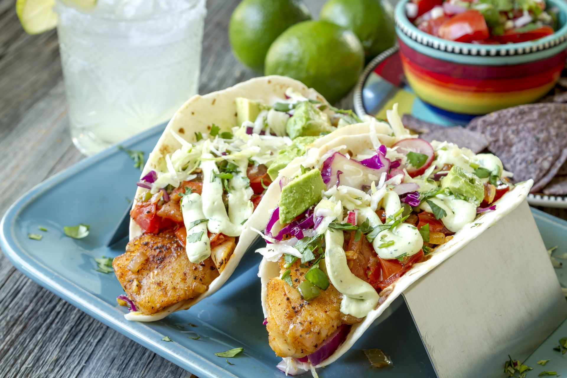 Two fix tacos with avocado, radishes, onions on a wood table with a drink and limes and guacamole in background