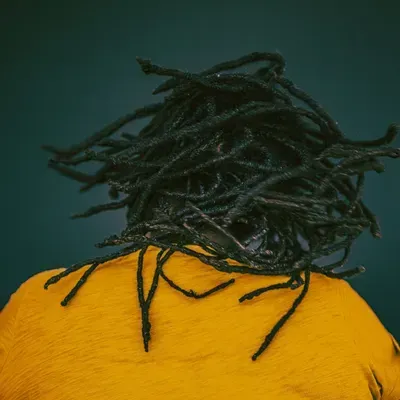 Person with black dreadlocks wearing a yellow shirt against a dark teal background