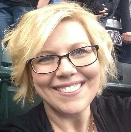 Woman with blonde hair and glasses smiles in a stadium, wearing a black top.