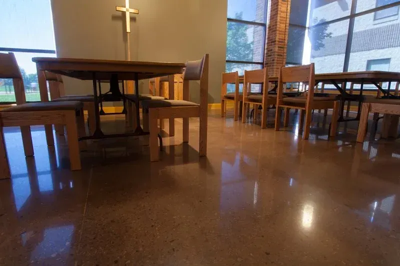 Empty church hall with wooden tables, chairs, and a cross on the wall, seen from floor level.