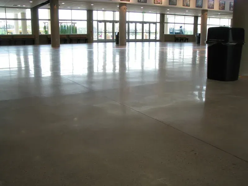 Empty indoor lobby with a polished concrete floor, glass entrance doors, and a trash can on the right.