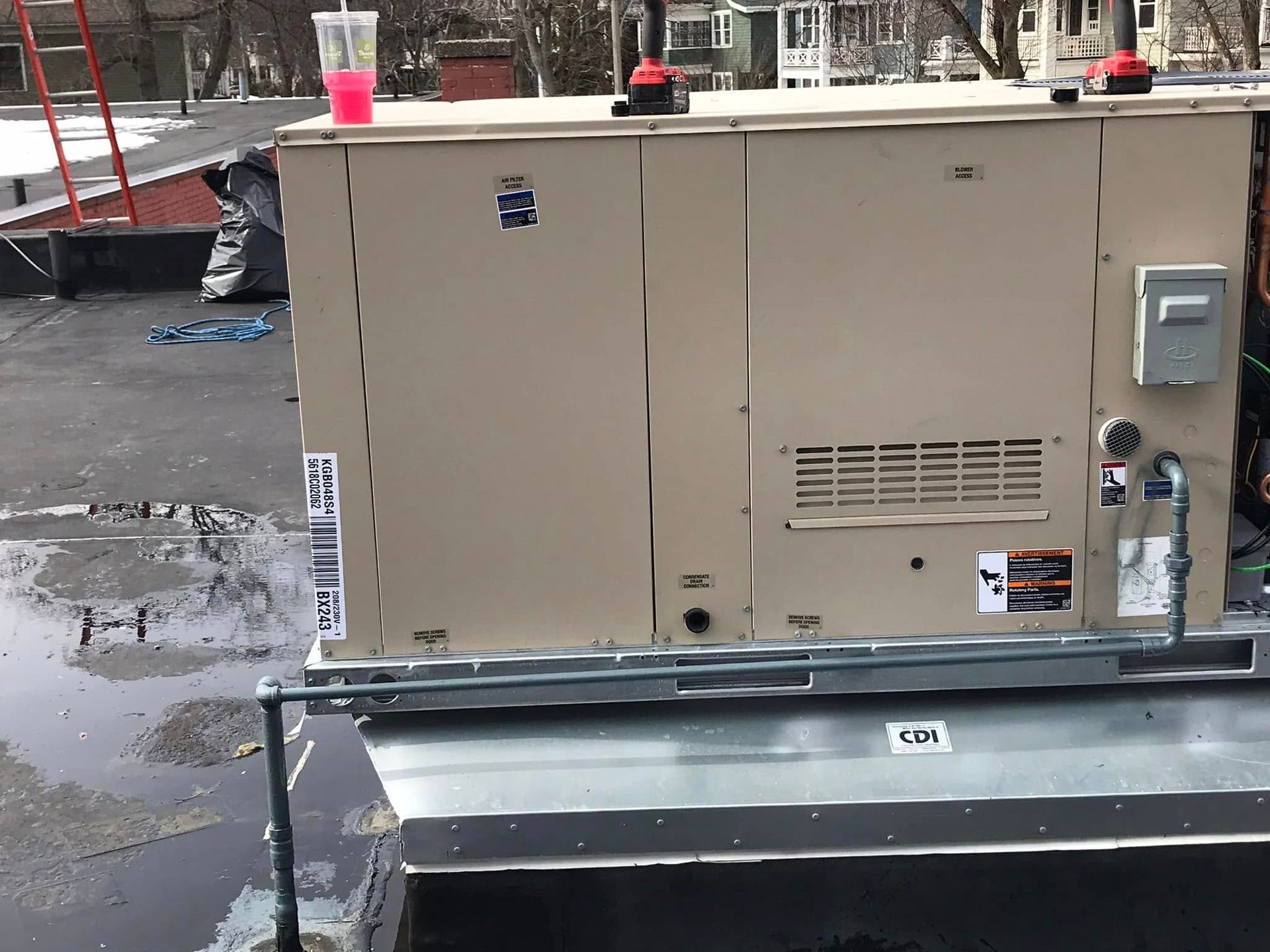 A beige industrial HVAC unit sits on a flat rooftop with a gas line connection, a red drink cup, and tools on top.
