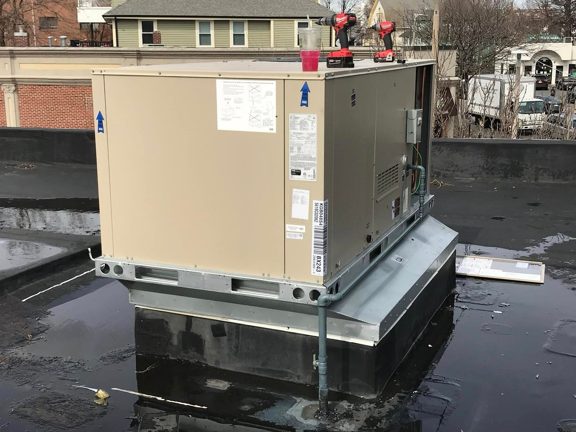 A tan rooftop HVAC unit sits on a metal curb on a wet, flat roof in an urban setting, with tools placed on top.