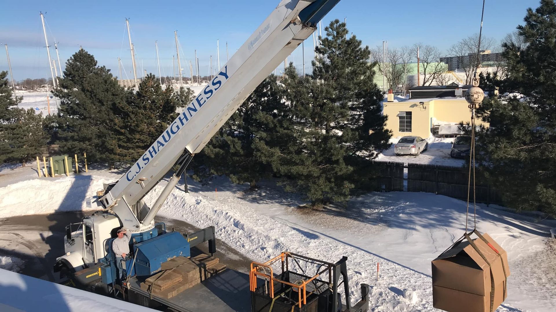 A crane truck lifts a large, brown, wrapped package in a snow-covered yard next to trees and buildings.
