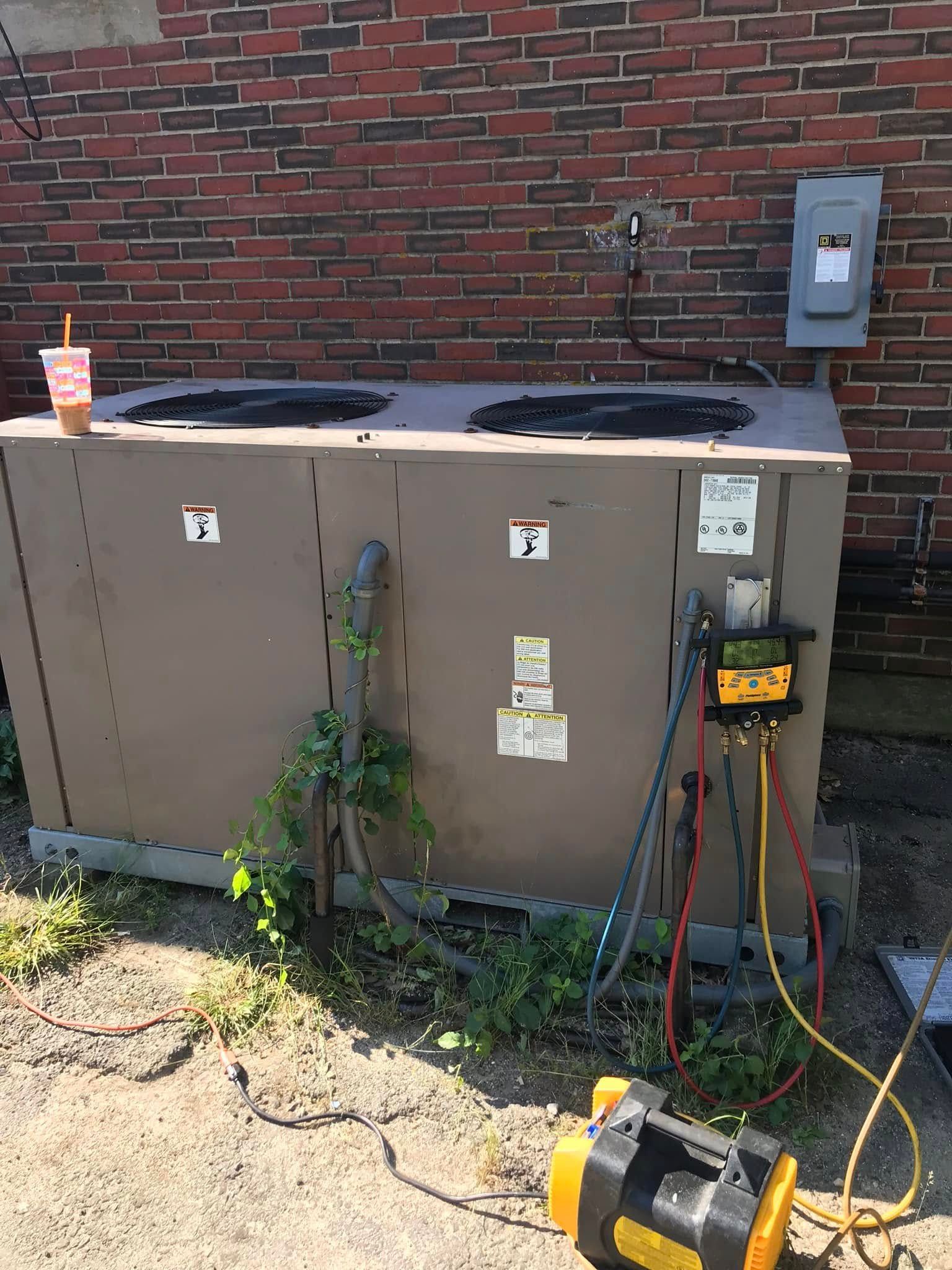 A tan industrial HVAC unit outdoors against a brick wall, connected to a technician's vacuum pump and gauge manifold.