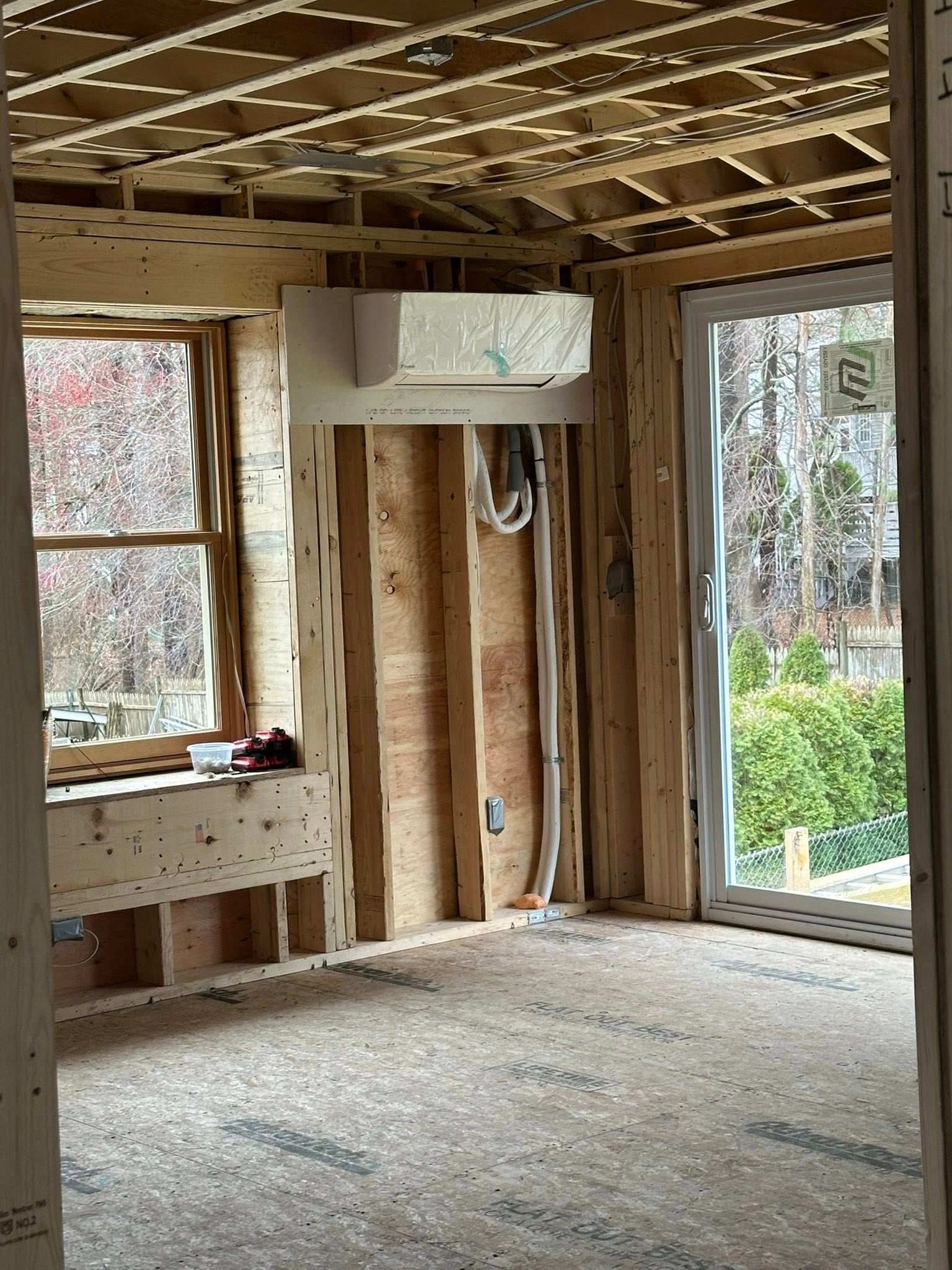 A wood-framed room under construction, showing a wall-mounted ductless mini-split unit, a window, and a sliding glass door.