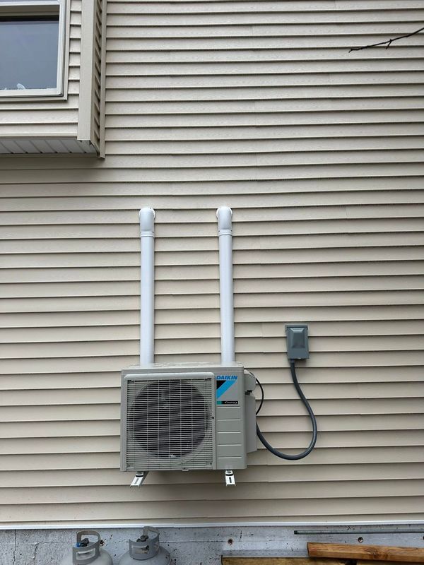 A Daikin ductless HVAC outdoor unit mounted on beige vinyl siding, with two vertical white conduits and a disconnect box.