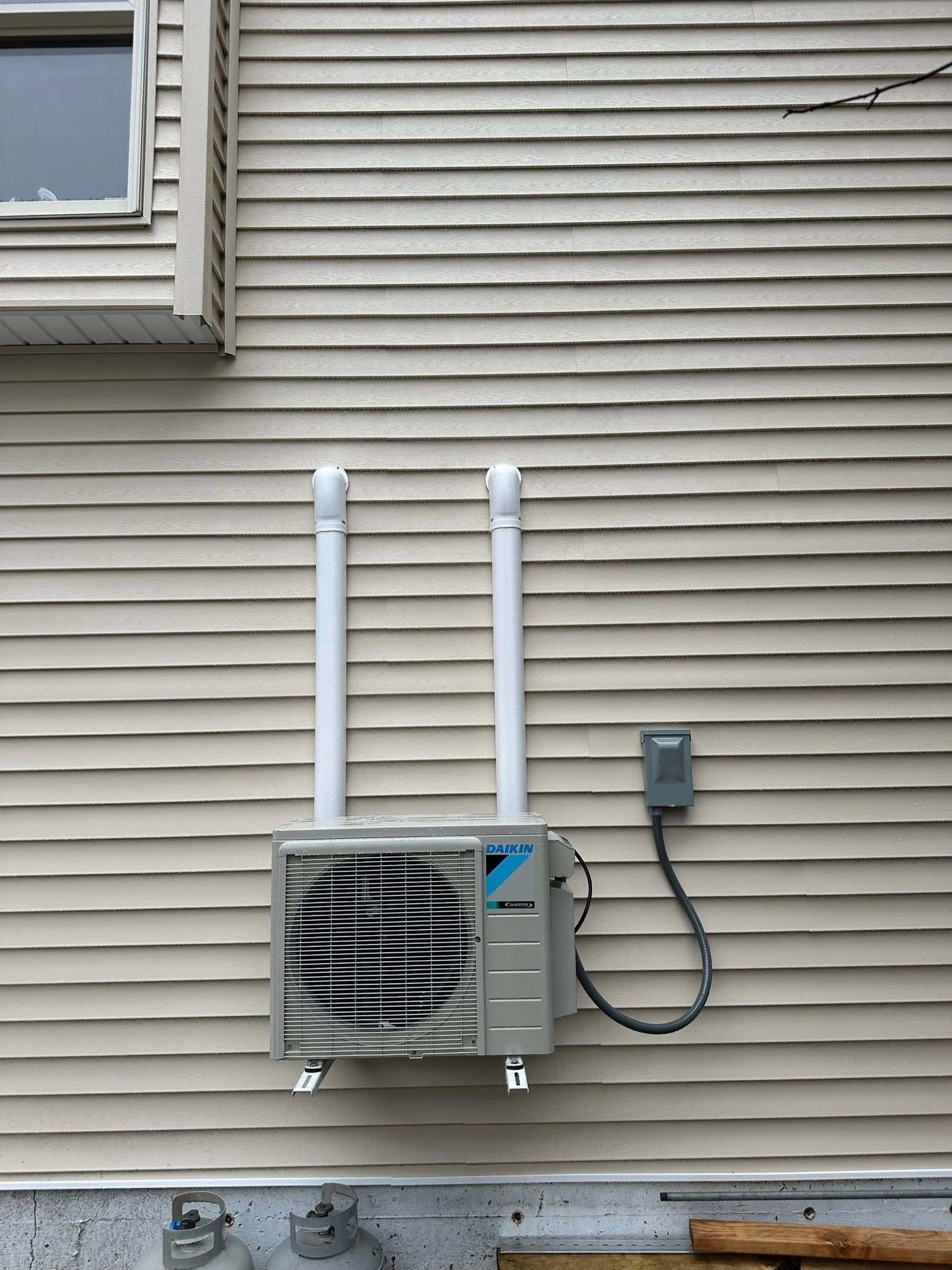 A Daikin outdoor HVAC unit mounted on tan siding, featuring two vertical white conduits and a gray electrical box.