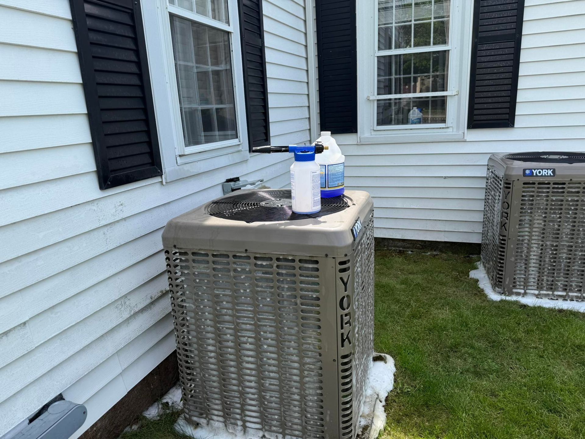 Two tan York air conditioning units sit outside a white-sided house, one with a spray bottle on top and suds on the grass.