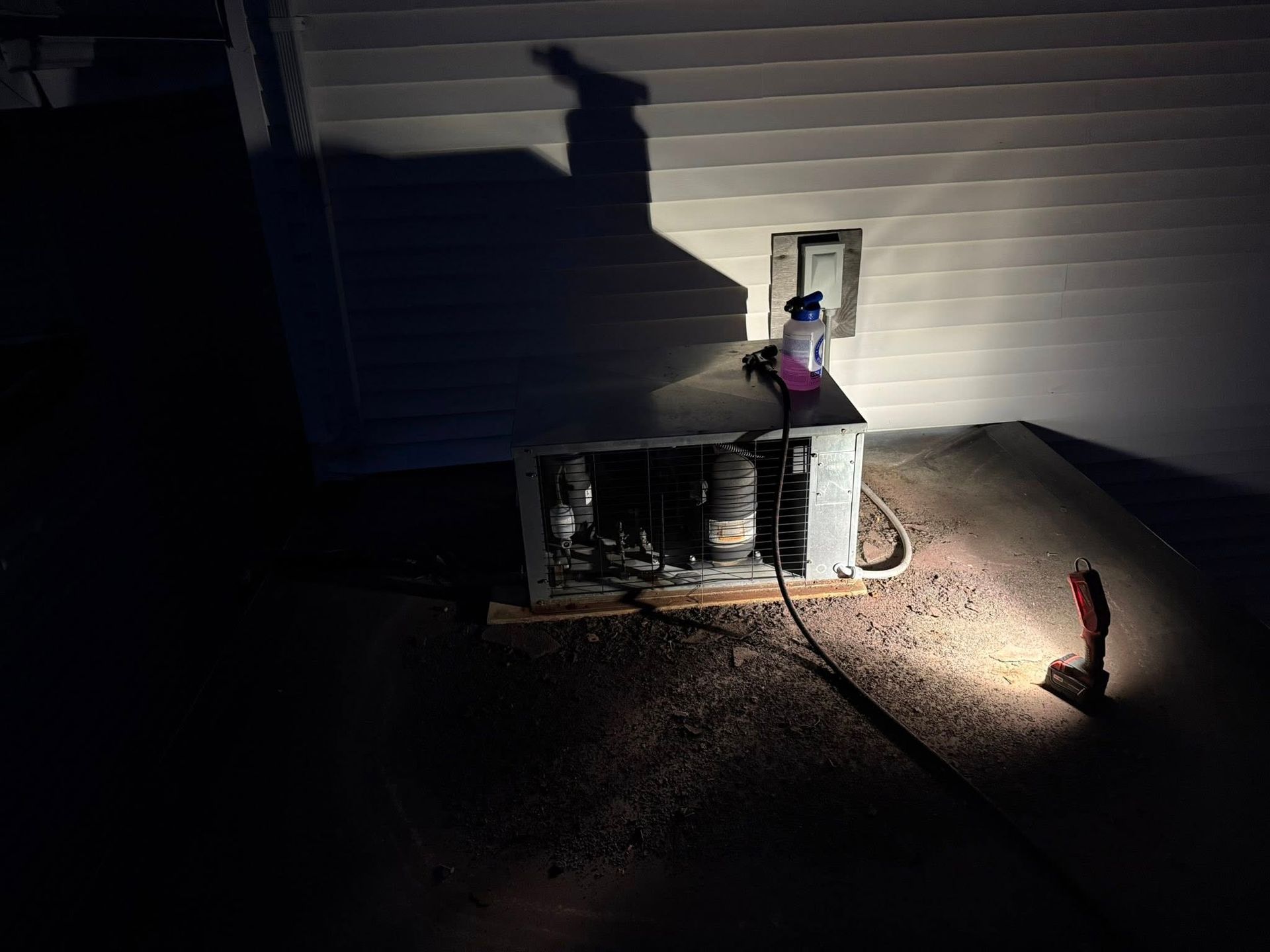 An air conditioning unit at night with an open maintenance panel and a light shining on the ground next to it.