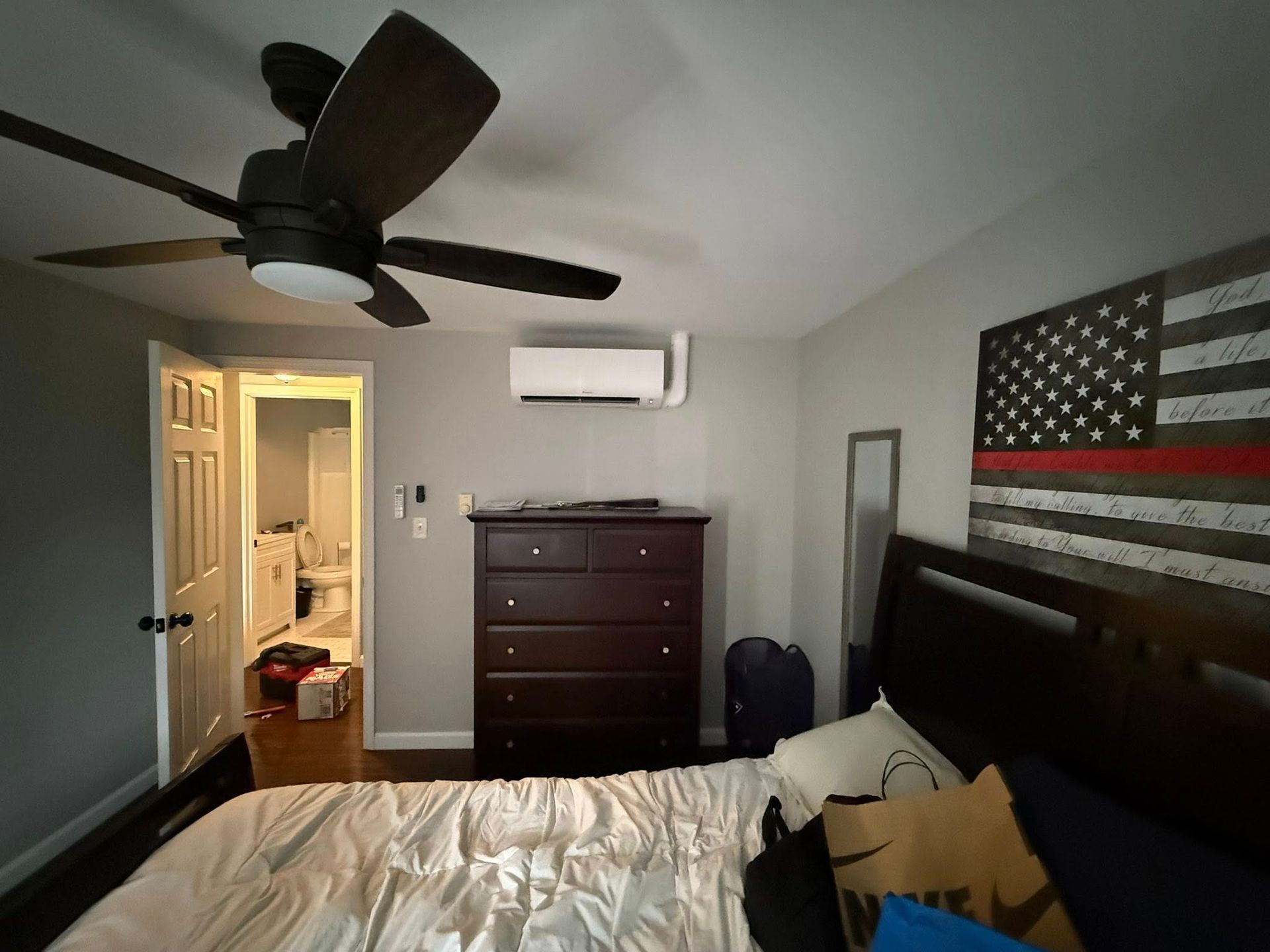 Bedroom featuring a dark dresser, ceiling fan, wall-mounted air conditioner, and a Thin Red Line flag above the bed.
