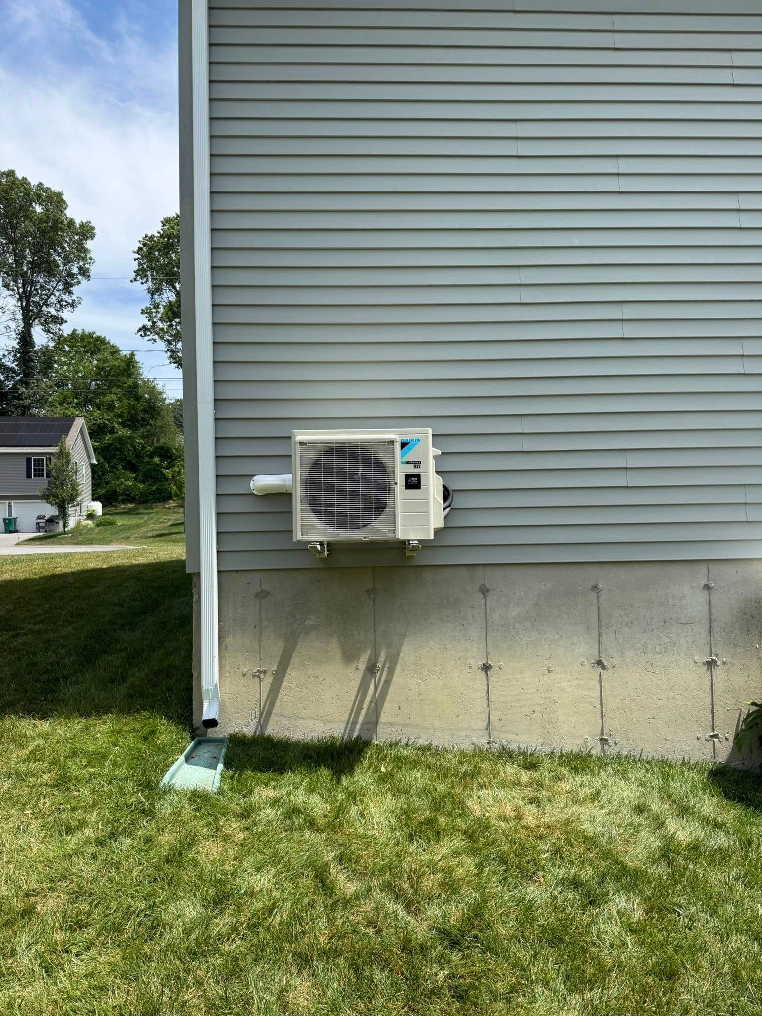 A white, wall-mounted Daikin mini-split outdoor AC unit attached to a grey-sided house above a concrete foundation.