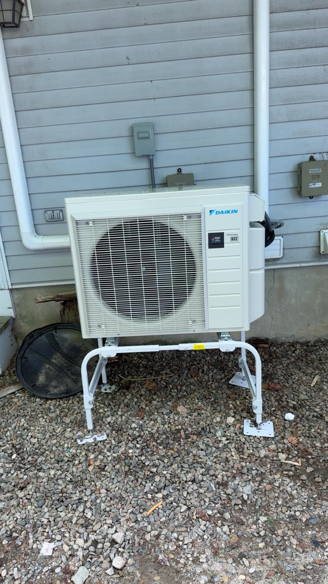 A Daikin outdoor HVAC unit mounted on a metal stand against a gray siding wall above a gravel ground surface.