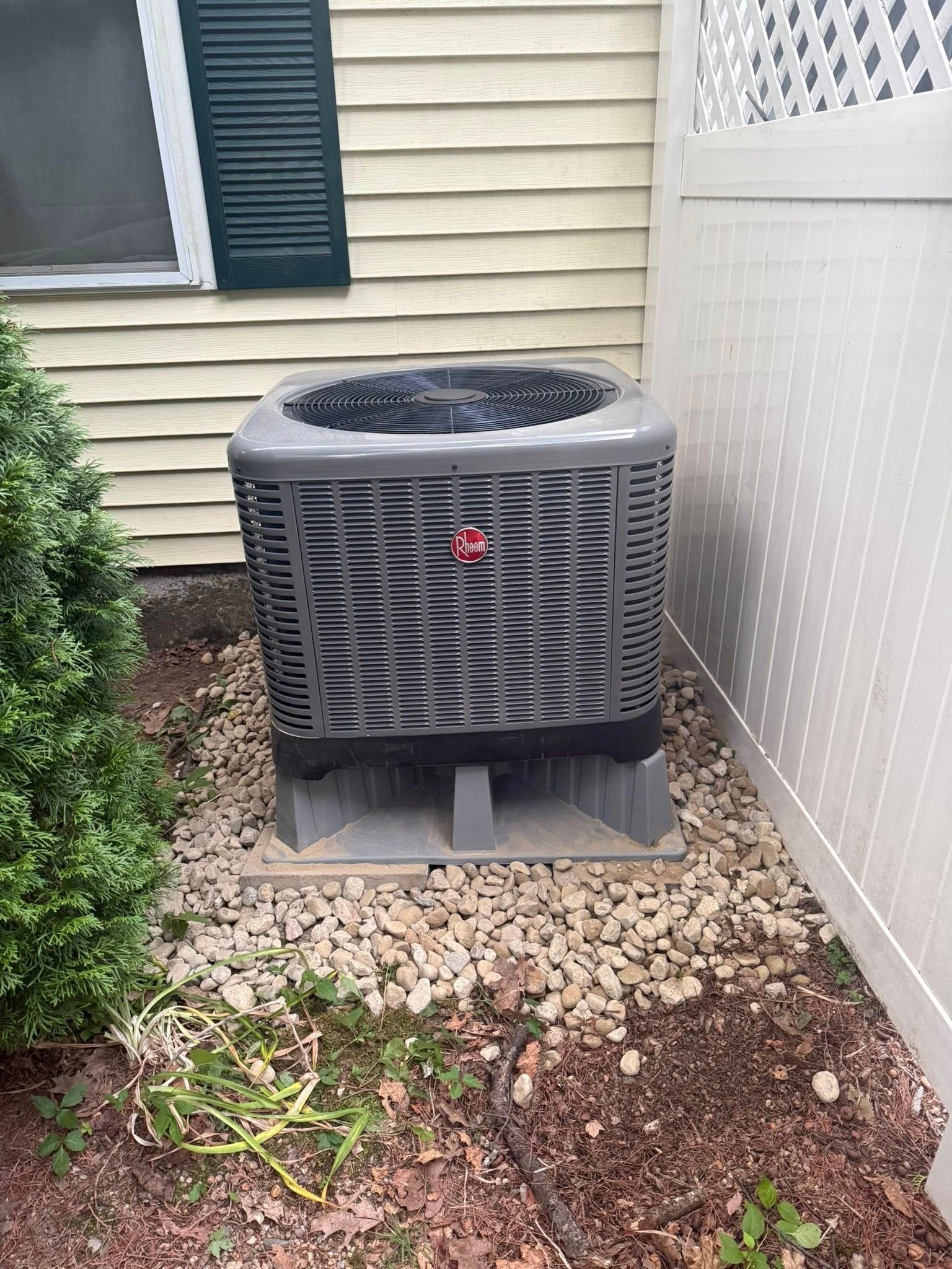 A gray outdoor air conditioning condenser unit sits on a gravel bed next to a house wall and a white vinyl fence.