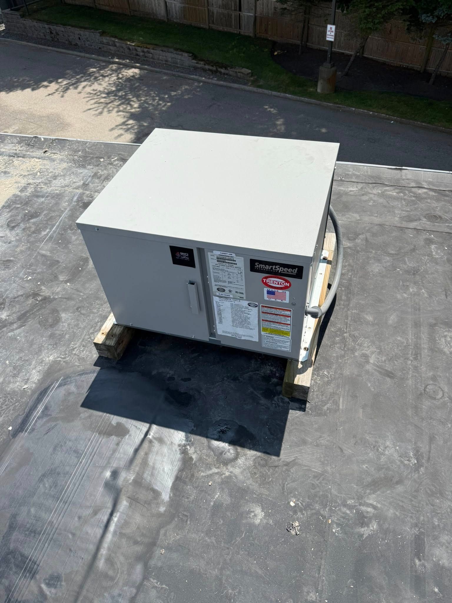 A gray electrical control box mounted on wooden blocks on a flat, dark-colored roof under bright, sunny conditions.