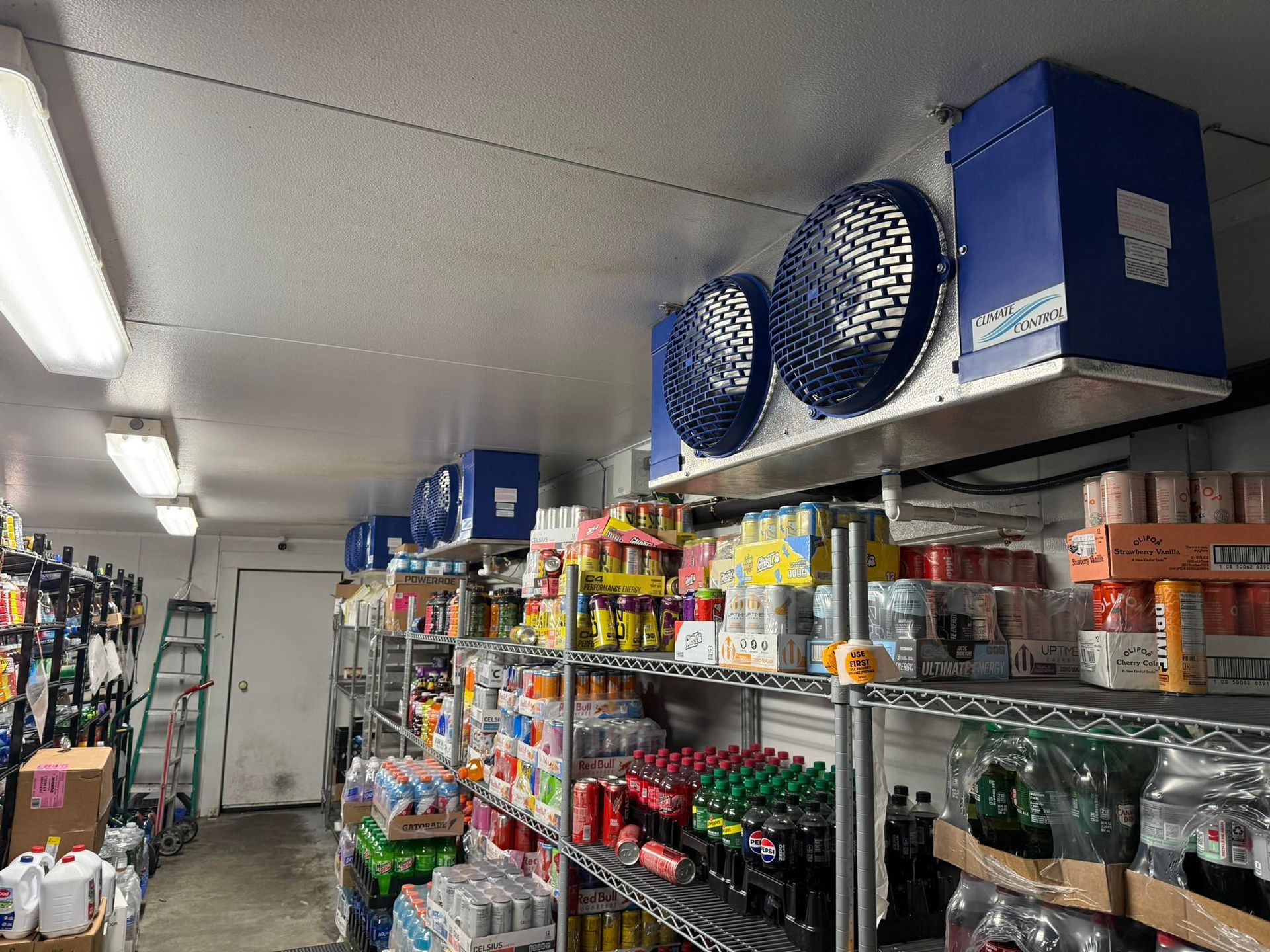 A well-stocked walk-in refrigerator featuring rows of metal shelves packed with drinks and groceries under blue fans.