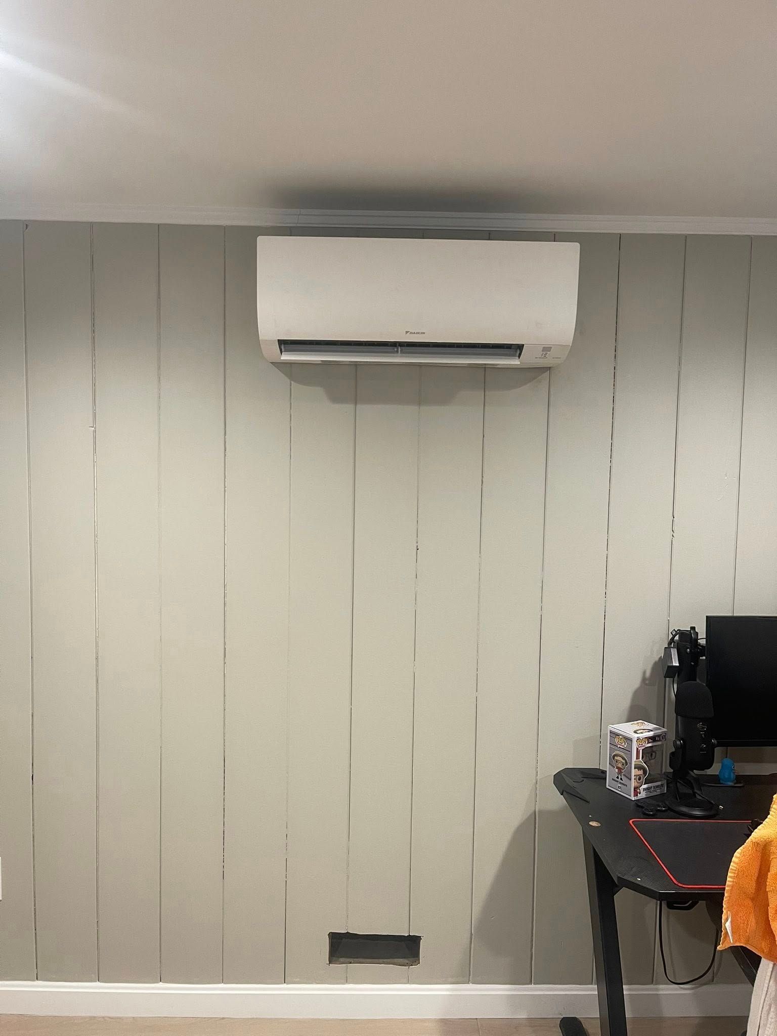 A white wall-mounted air conditioner centered on a light gray wall with vertical metallic stripes, above a computer desk.