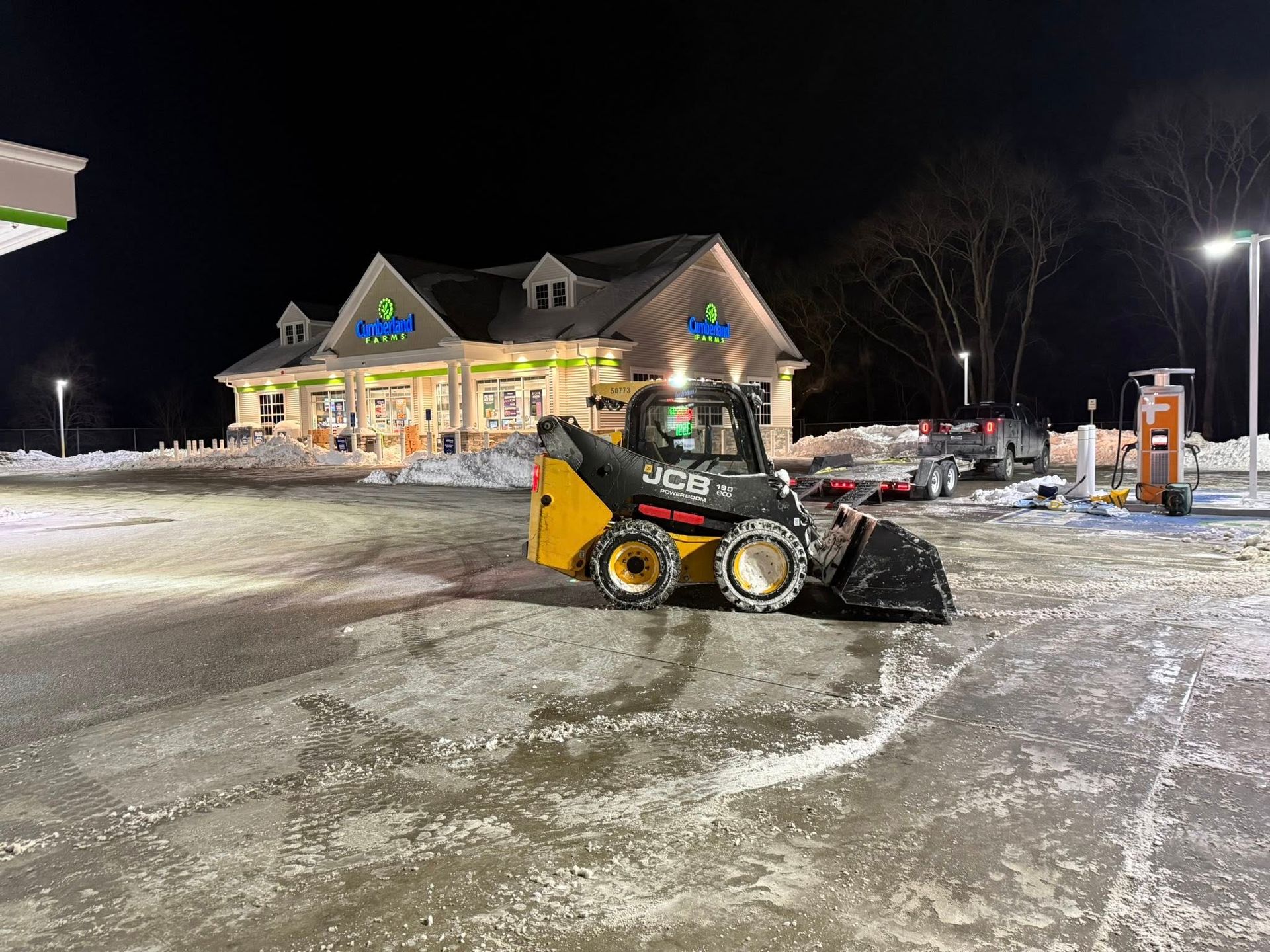 A yellow and black skid-steer loader plows snow in a parking lot at night in front of a brightly lit convenience store.