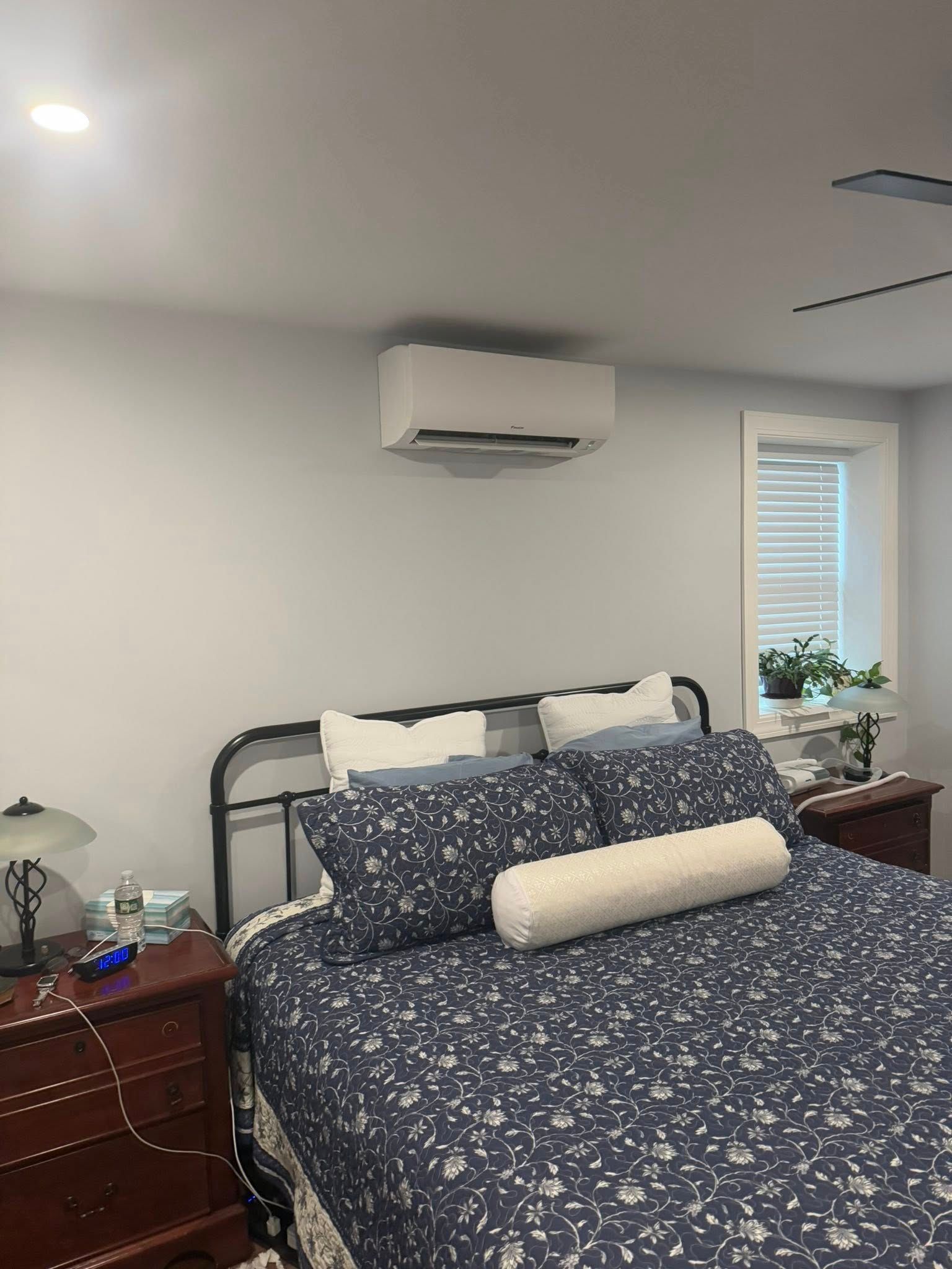 A bedroom featuring a bed with blue patterned bedding and a wall-mounted AC unit above the headboard next to a window.