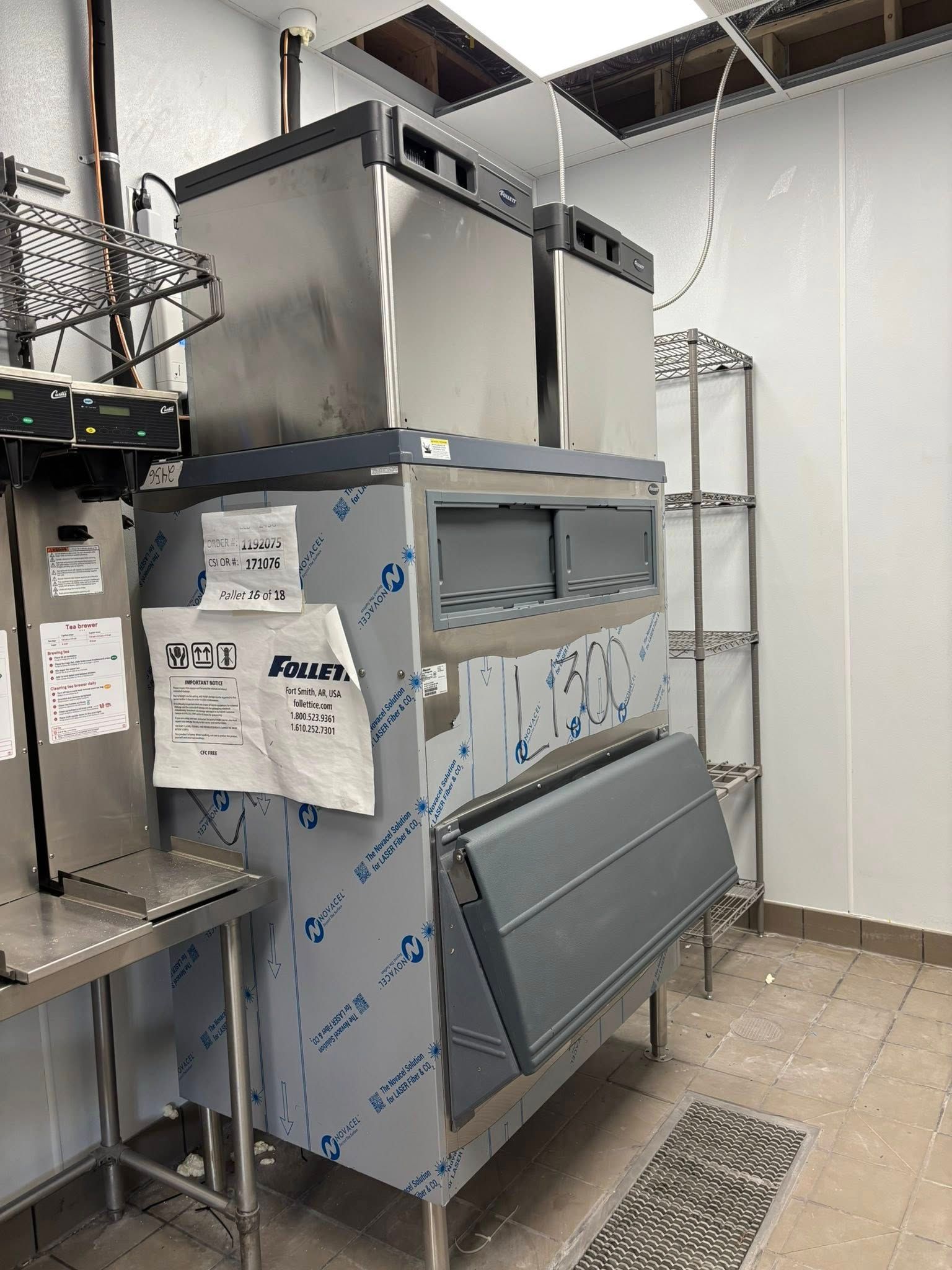 A stainless steel ice machine and bin stand in a utility room with white walls, tiled floors, and exposed ceiling panels.