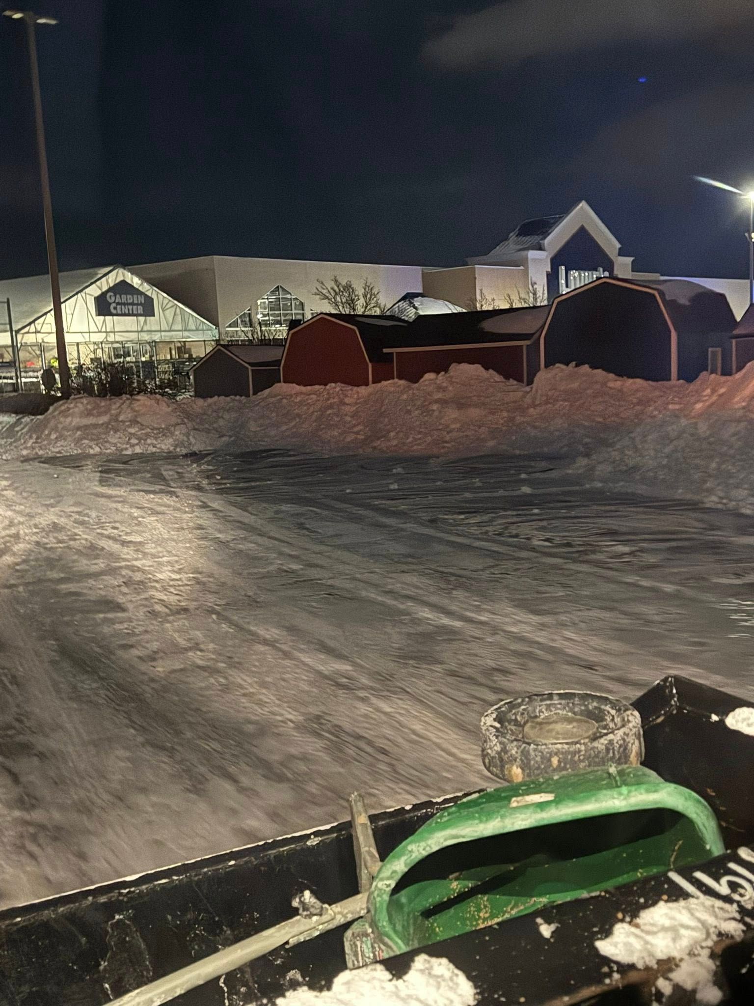 A snowy parking lot at night with red sheds in the background, viewed from the perspective of a snowplow operator.