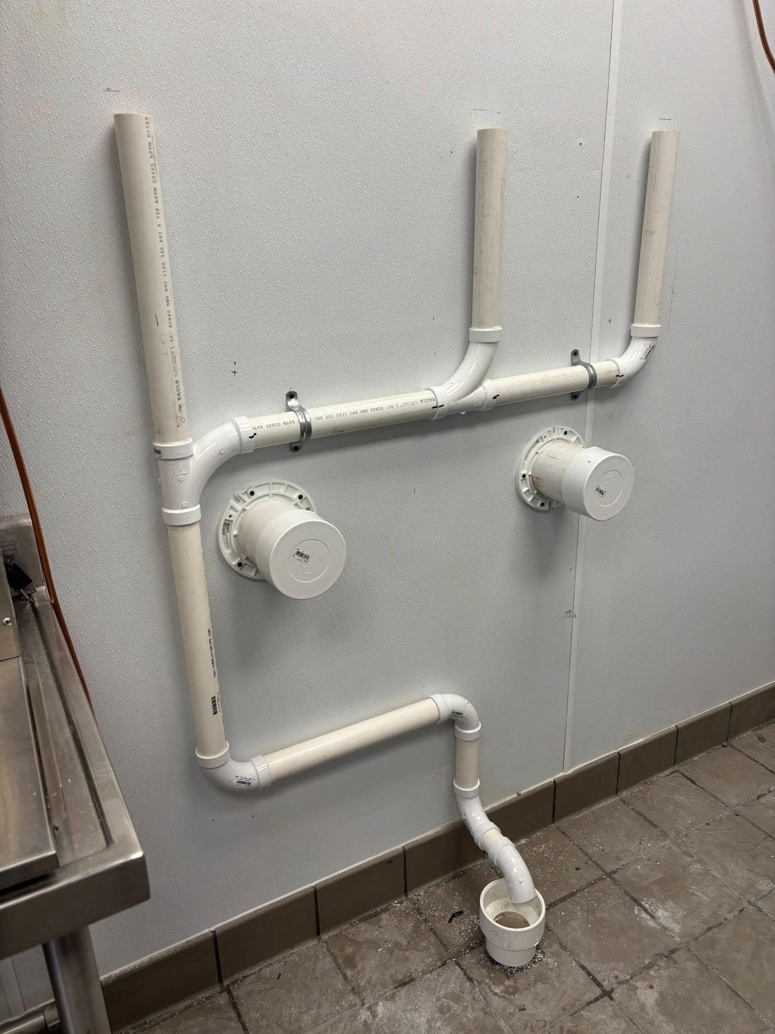 White PVC plumbing pipes mounted on a wall above a floor drain in a commercial kitchen setting.