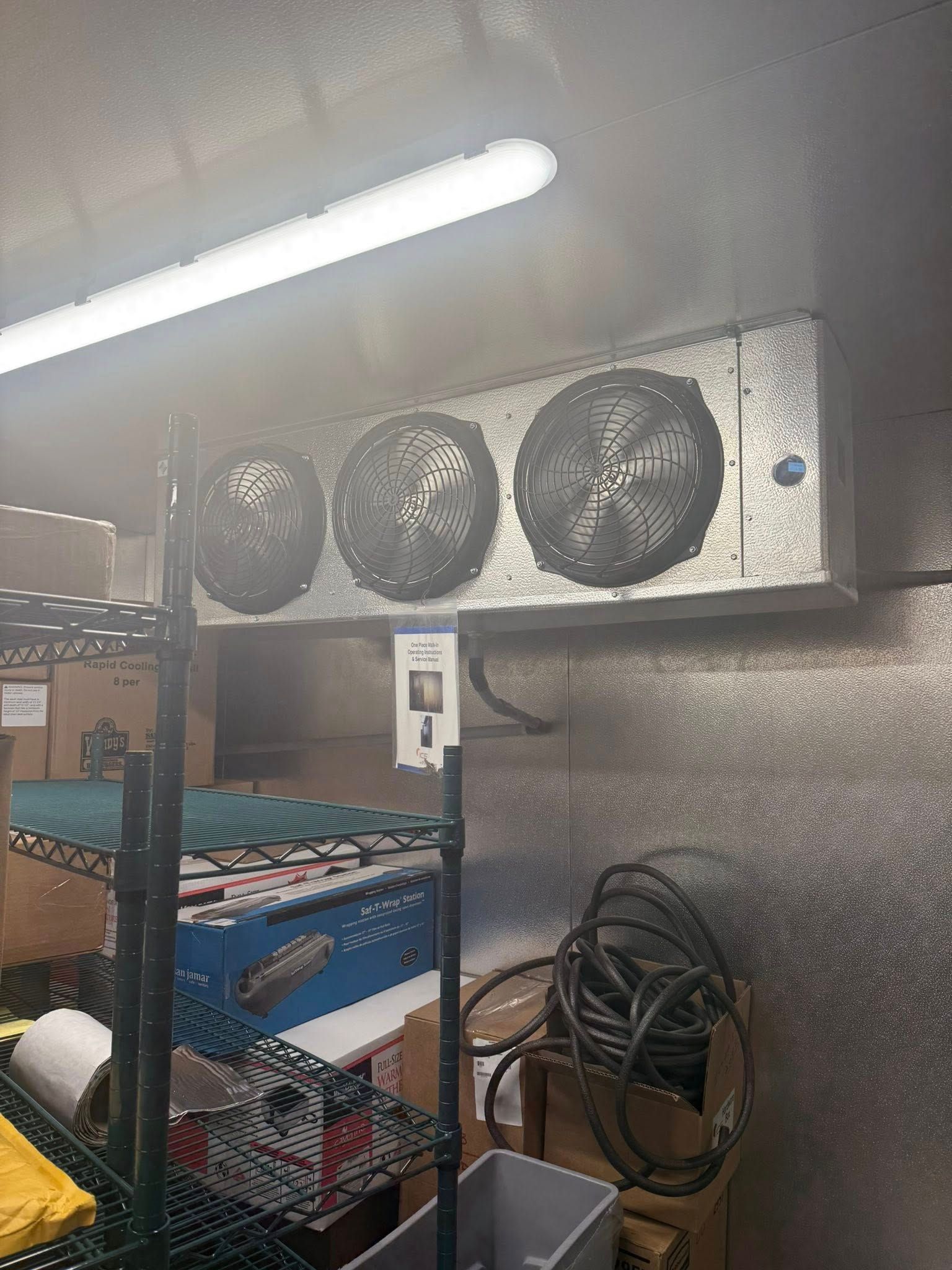 Industrial refrigeration unit with three fans mounted on a metal wall inside a walk-in cooler storage area.