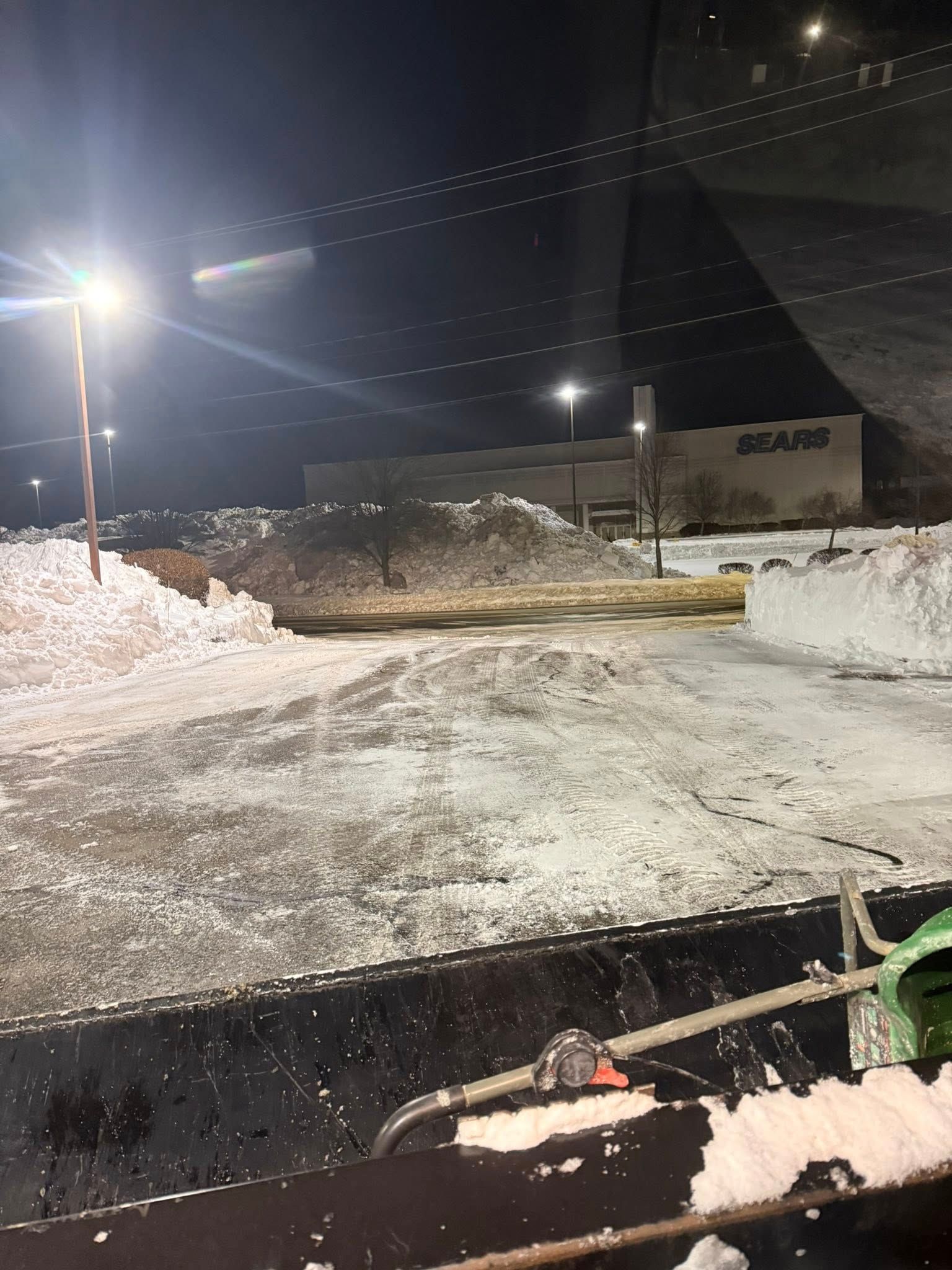 A view from a plow truck cabin at night, showing a snow-covered parking lot with large piles of snow and a Sears store.