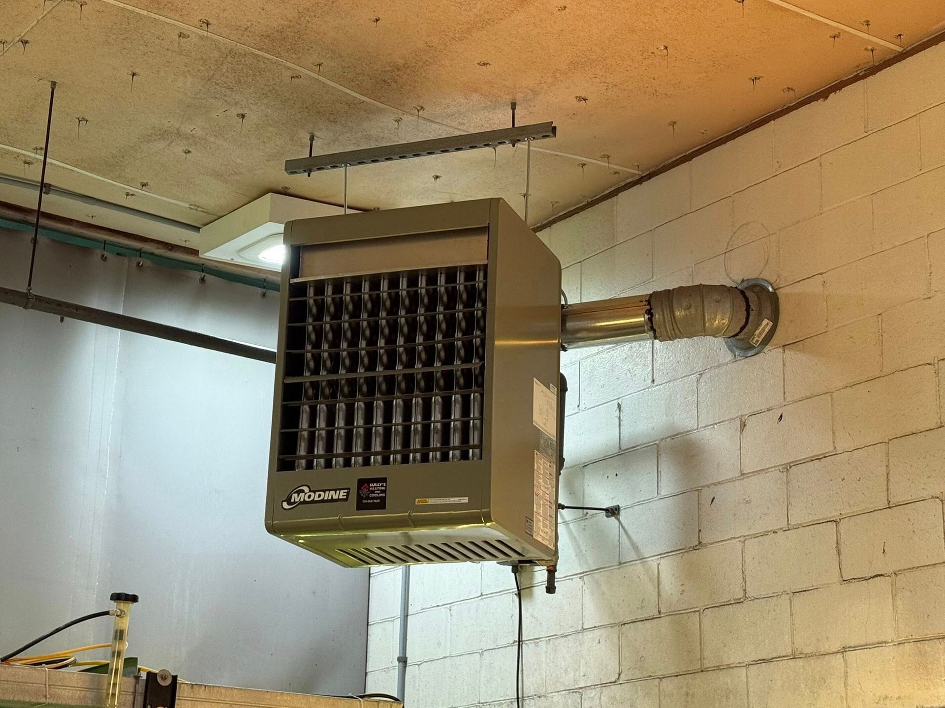 A tan industrial heater suspended from a ceiling by metal rods, with a vent pipe connected to a white brick wall.