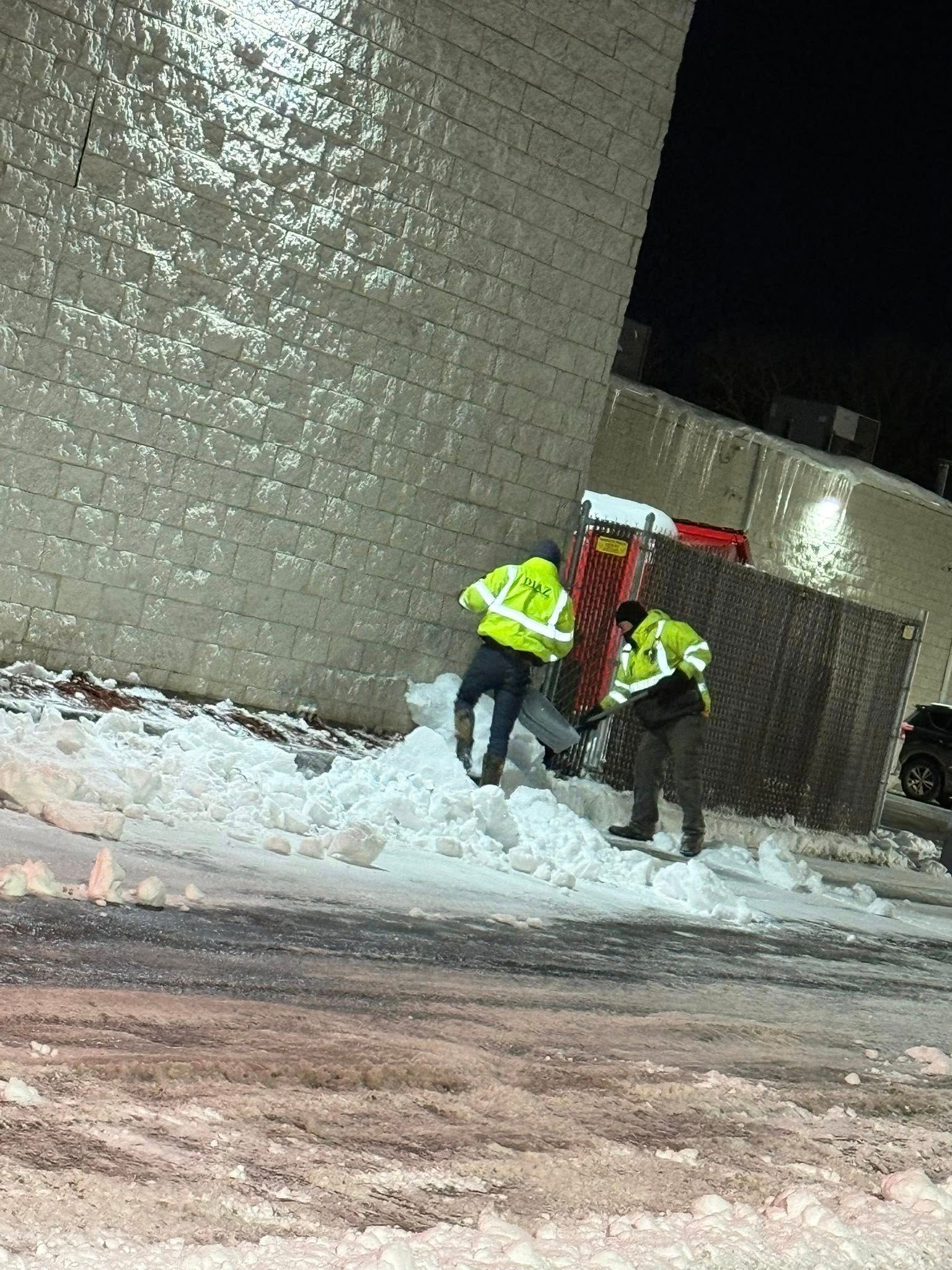 Two people in high-visibility yellow jackets clear snow from near a building wall at night.