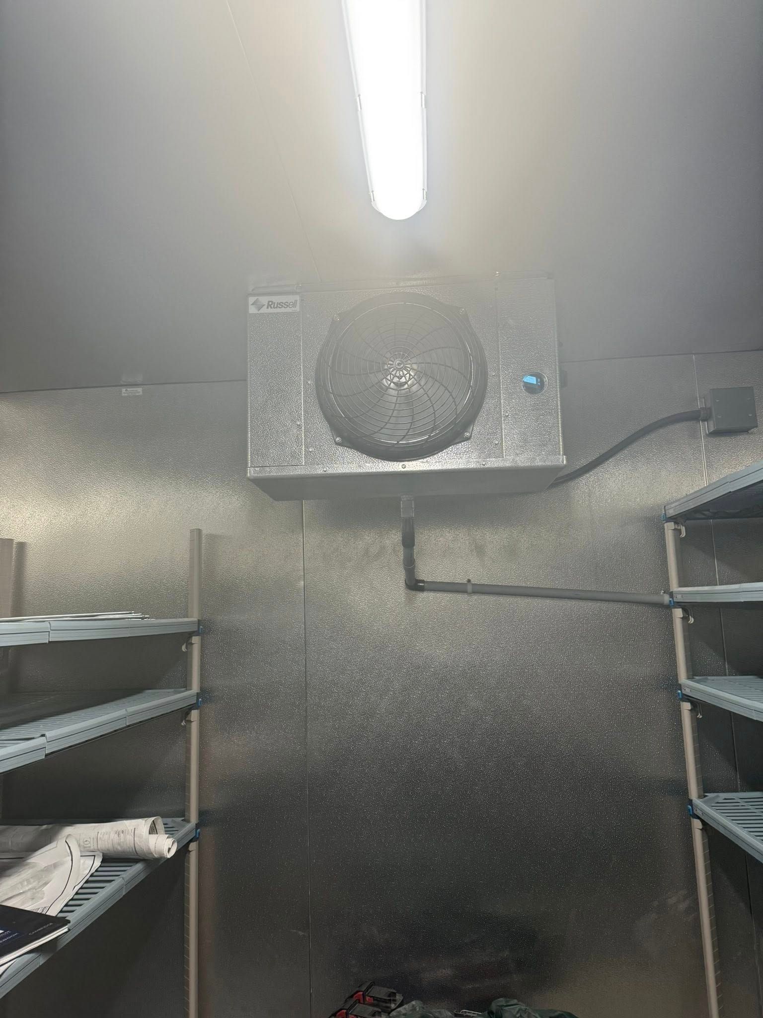 A walk-in refrigerator unit with a cooling fan, mounted on a textured, metallic wall between two storage shelves.