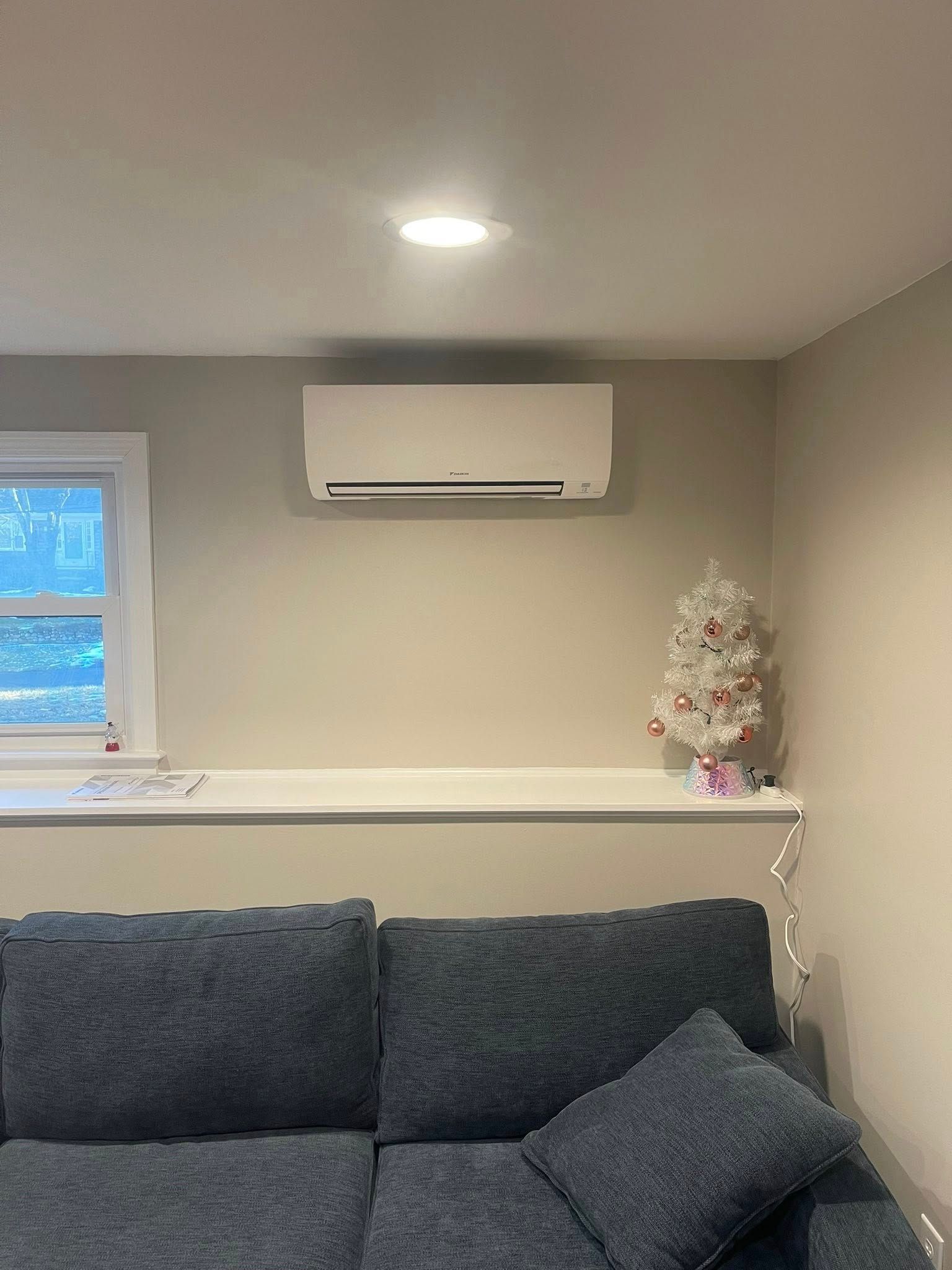 A gray sofa sits below a wall-mounted white air conditioner and a ledge featuring a small, decorated miniature tree.