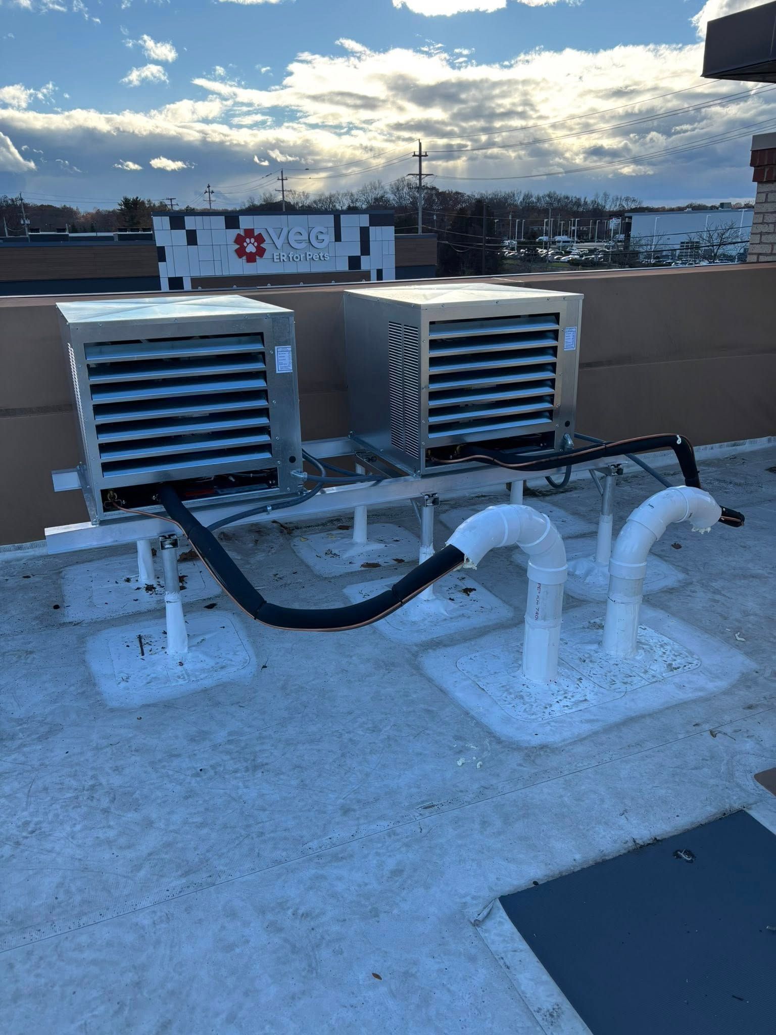 Two silver HVAC units sit on a rooftop on elevated metal stands, connected by black insulated lines and white conduits.