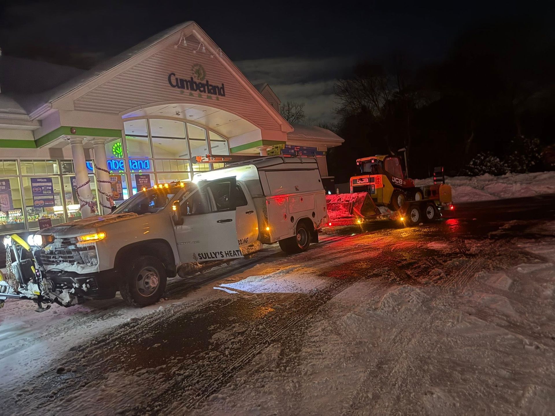 A white service truck with a snow plow and a yellow skid steer parked in front of a Cumberland Farms at night.