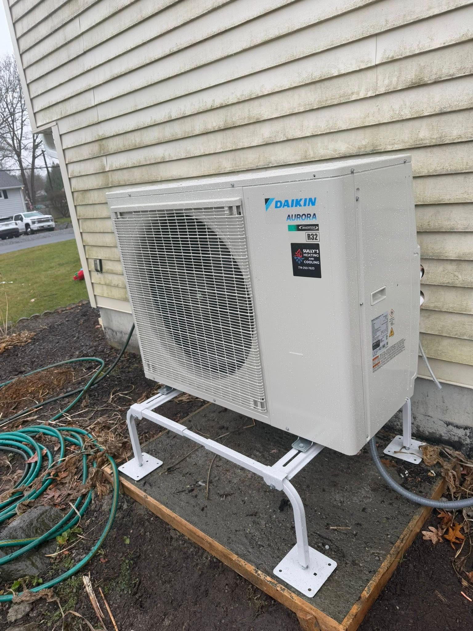 A Daikin outdoor HVAC unit sits on a white metal stand on a concrete pad next to the tan vinyl siding of a house.