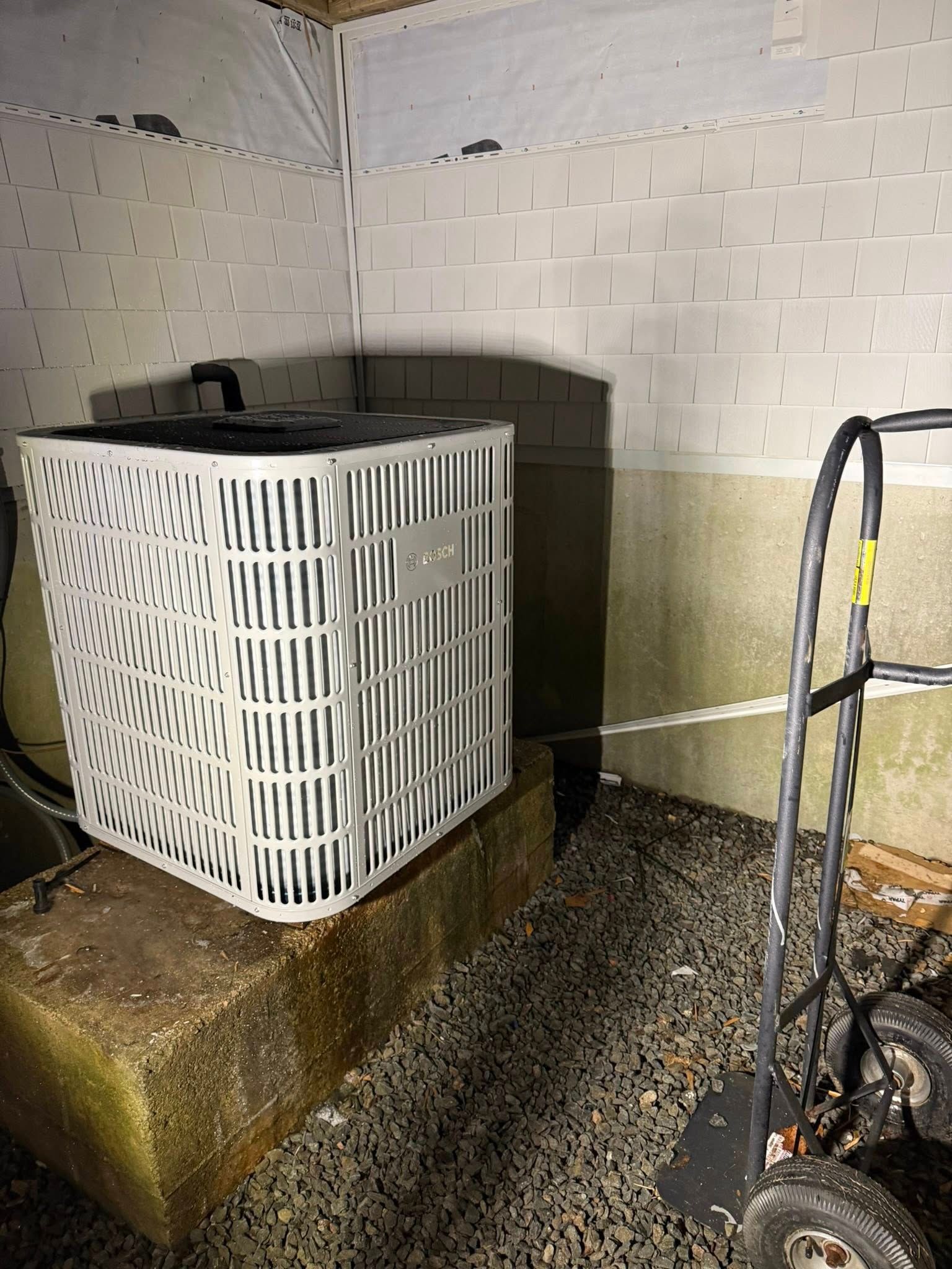 A white HVAC unit sits on a concrete block against a white cinder block wall in a gravel area next to a hand truck.