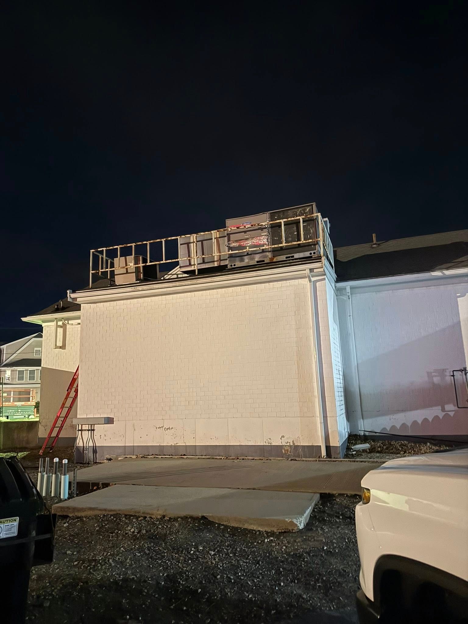 A construction site at night showing a building's white exterior wall with exposed framing on the roof and a red ladder.