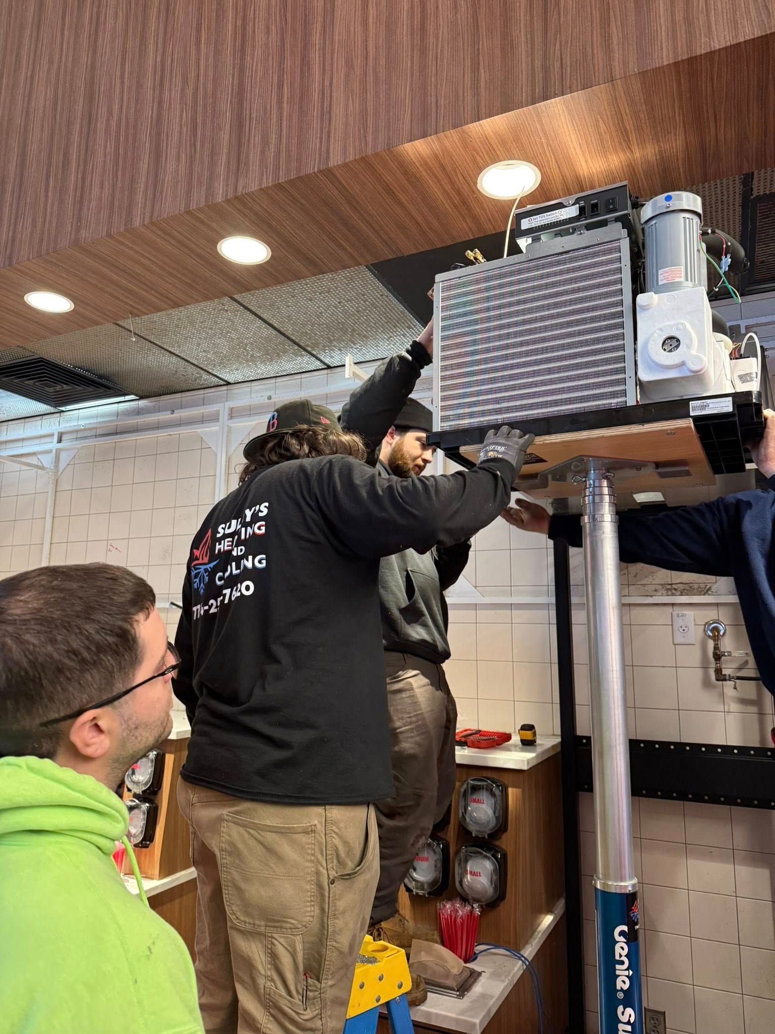 Technicians work together to install a large industrial HVAC unit suspended from the ceiling in a commercial space.