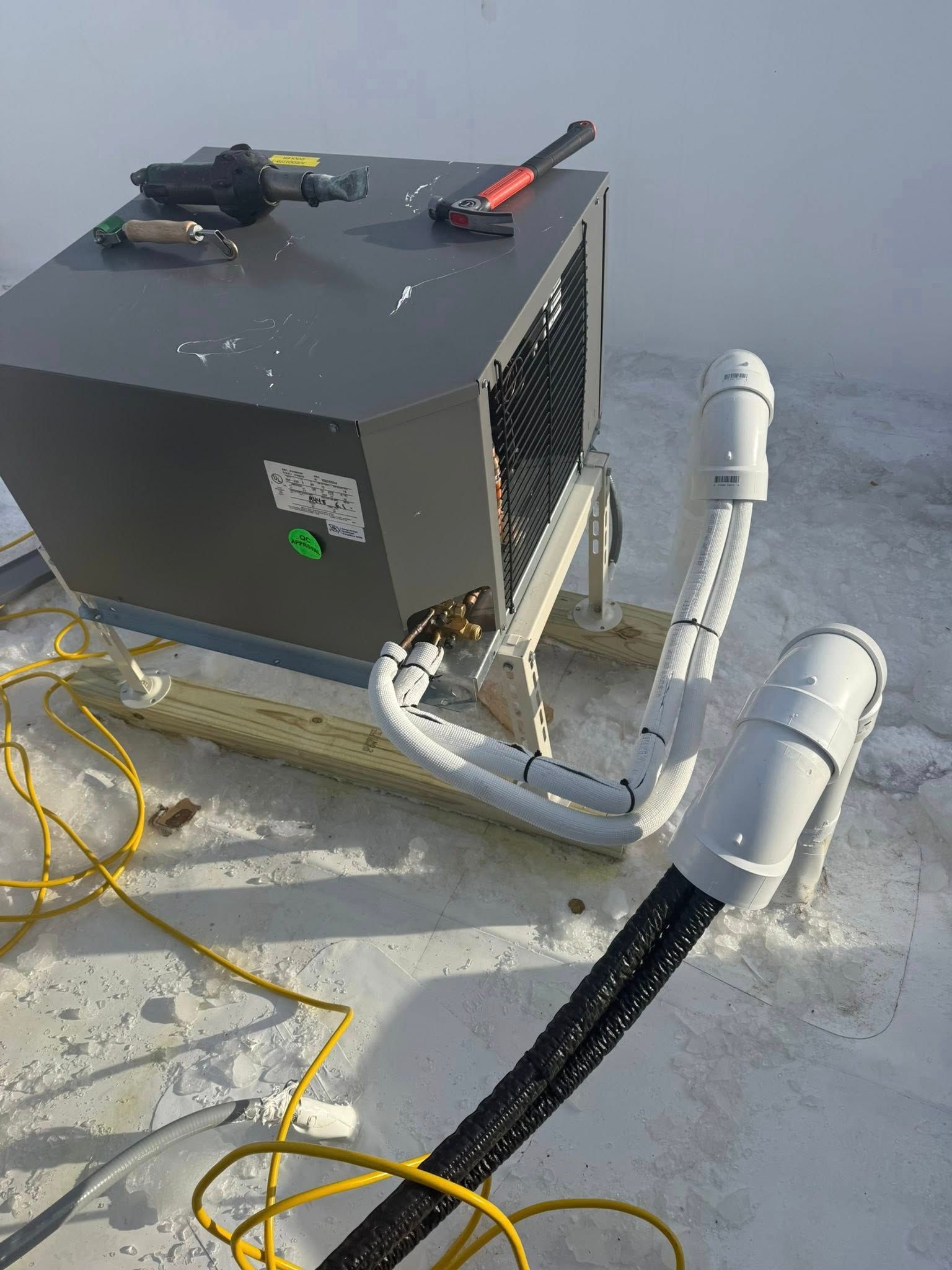 A grey HVAC unit on a white roof with white PVC pipes connecting to a black cable, tools resting on top.
