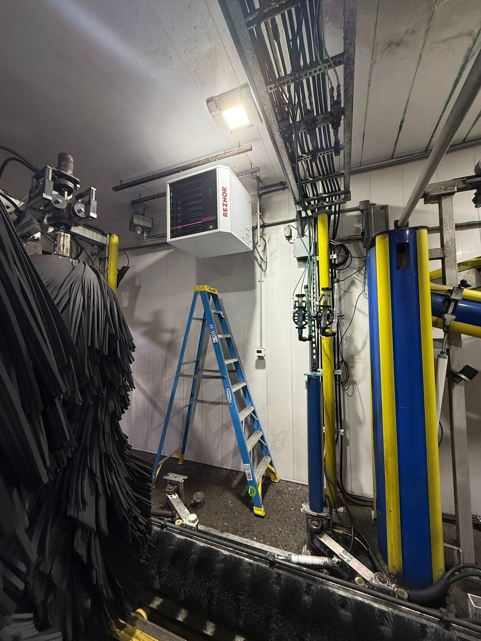 A stepladder sits in a car wash bay under a white industrial heater and overhead metal conduits.