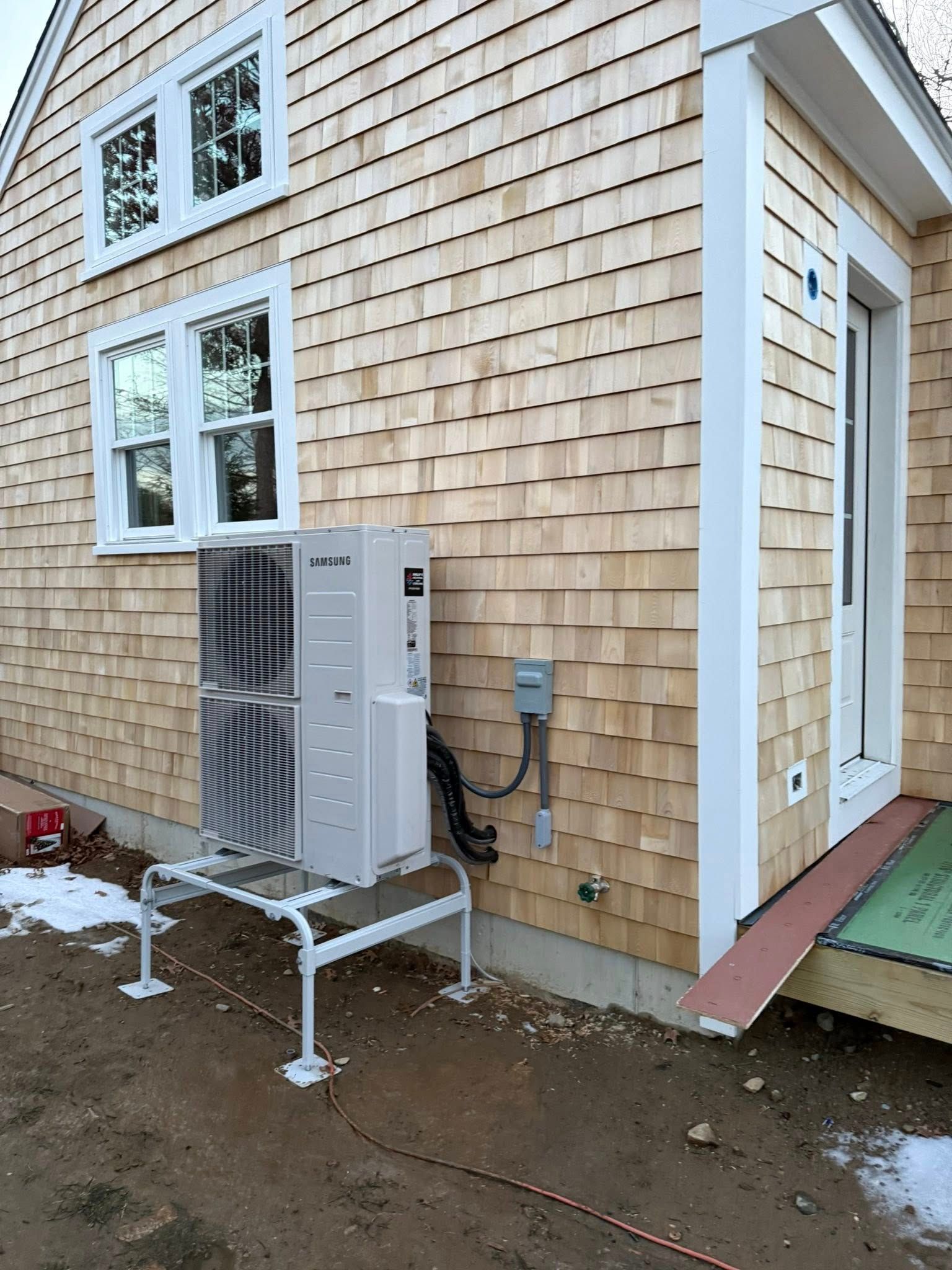 A mini-split heat pump unit stands on a white metal frame outside a house clad in light-colored wooden shingles.