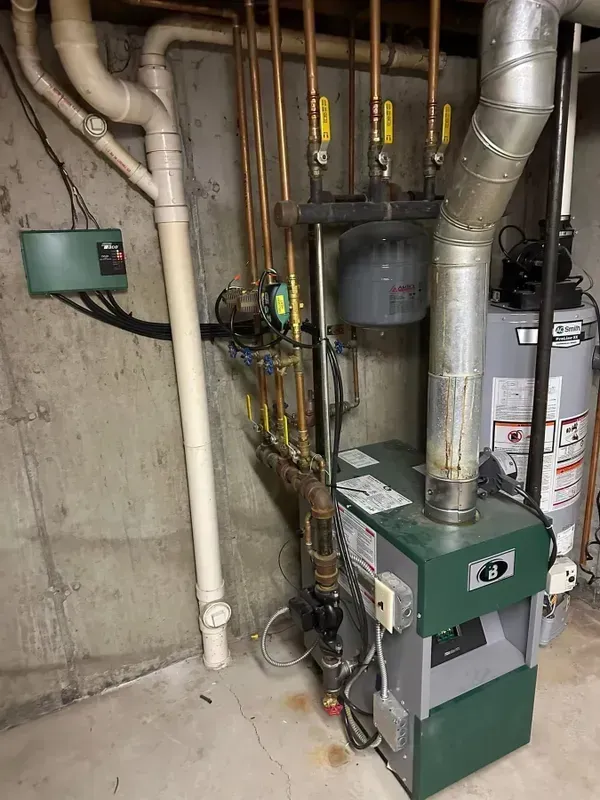 A home basement corner featuring a green boiler, metal exhaust vent, copper piping, and a nearby water heater.