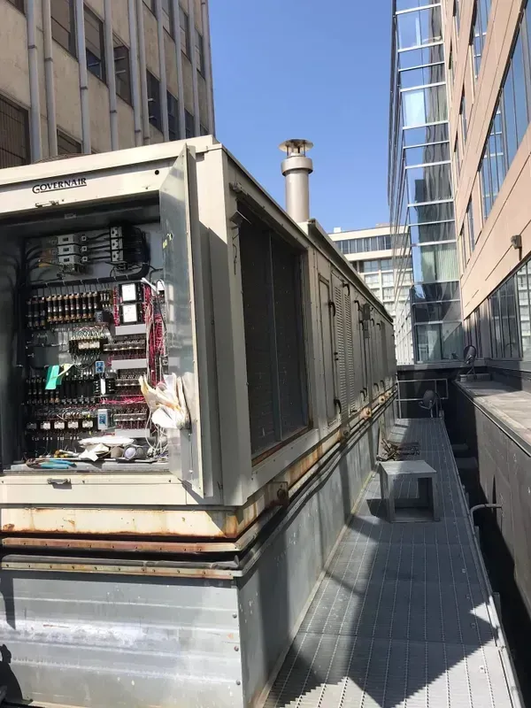 An open industrial HVAC unit on a rooftop walkway between two large buildings on a sunny day.