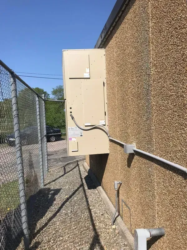 A tan, wall-mounted utility unit attached to a pebble-dash exterior wall next to a chain-link fence.