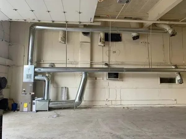 HVAC system with metal ductwork mounted on a light-colored industrial wall inside an unfinished room.