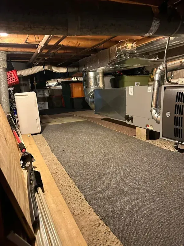 A low-ceilinged basement utility area with an HVAC unit, metal ductwork, and a gravel floor with a central mat.