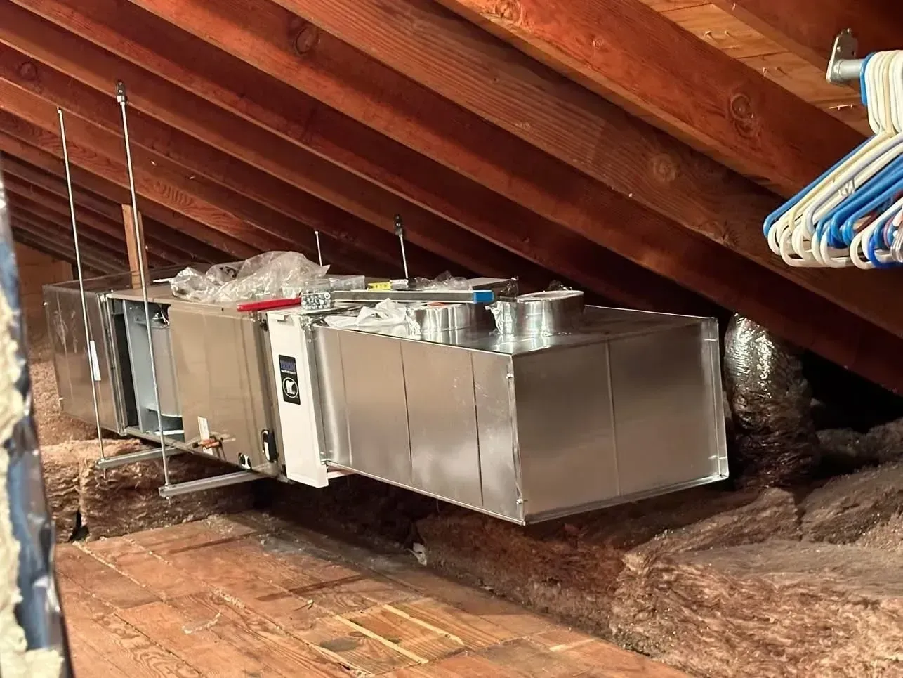 A metal HVAC air handler unit suspended in an attic space with wooden rafters and fiberglass insulation below.