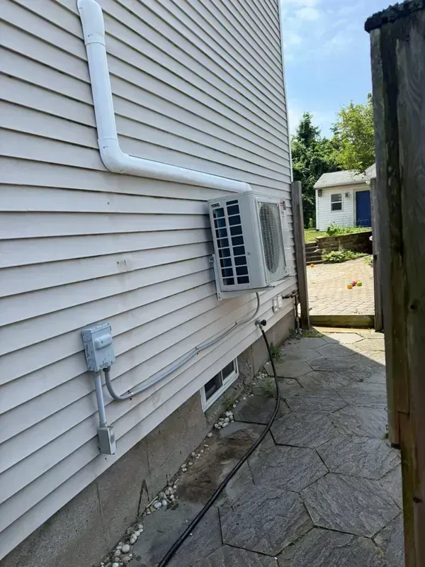 A mini-split air conditioner unit is mounted on the light-colored vinyl siding of a house exterior near a stone path.