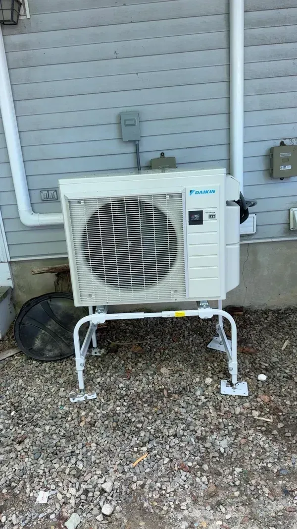 A white Daikin outdoor HVAC unit on a metal stand mounted against a gray vinyl-sided exterior wall.