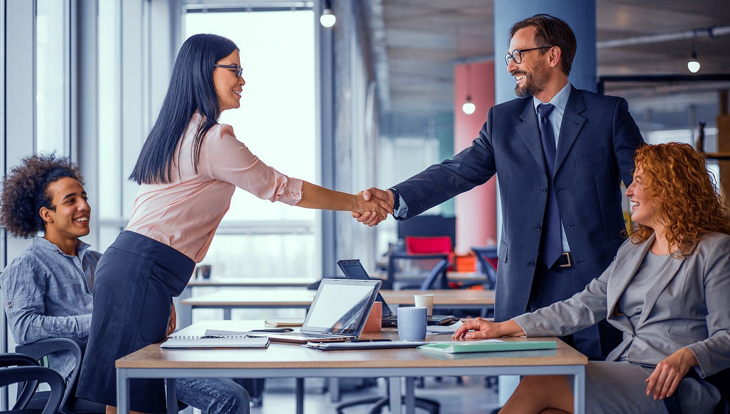 Two coworkers shaking hands in a modern office meeting room while others sit nearby with laptops